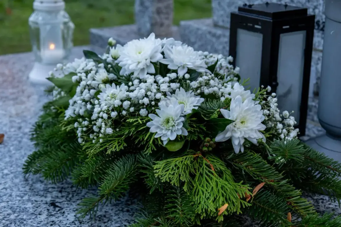 Białe wiązanki na grób, ozdobione zielenią i drobnymi kwiatami, spoczywają na kamiennym nagrobku obok znicza i latarenki.