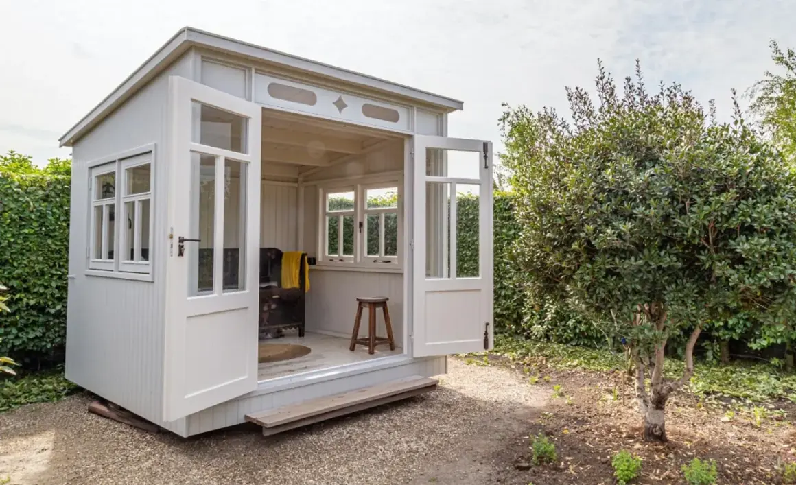 Eine charmante Gartenlaube mit offenen Türen, bereit, mit gemütlichen Möbeln eingerichtet zu werden. Ein Hocker und eine Decke deuten auf eine entspannte Atmosphäre hin.