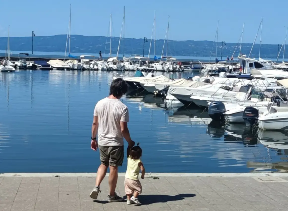 Padre e figlia passeggiano sul lungolago di Bolsena, con barche ormeggiate sullo sfondo.