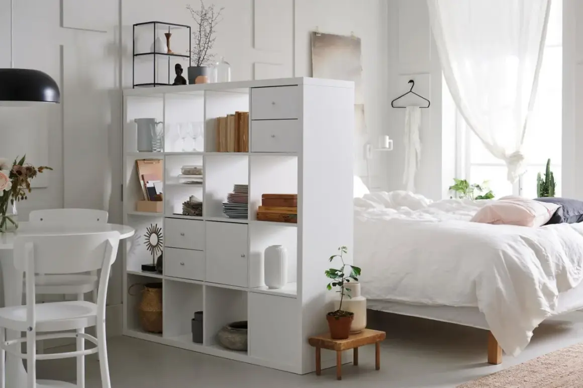 Ein heller Raum mit einem weißen Regal, das als Trenner dient. Links ein Essbereich, rechts ein Bett – ein cleverer wohn und schlafzimmer in einem raum.