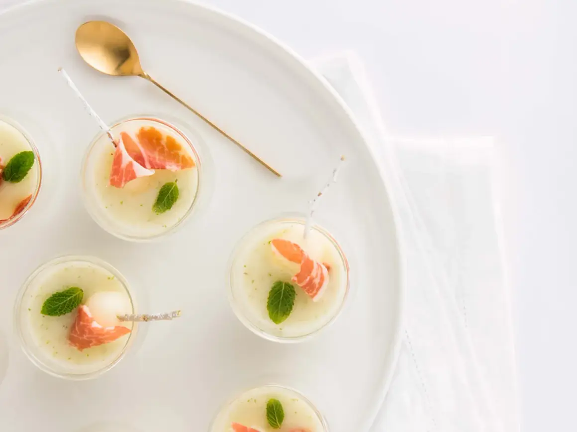 Delicioso chupito de melón con jamón serrano y menta, servido en copas pequeñas con una cuchara dorada.