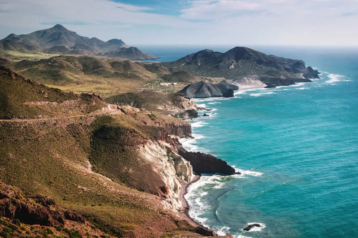 Costa escarpada cerca de Níjar, con el mar turquesa rompiendo en acantilados blancos y rojizos. Montañas verdes y una carretera serpentean en la distancia.