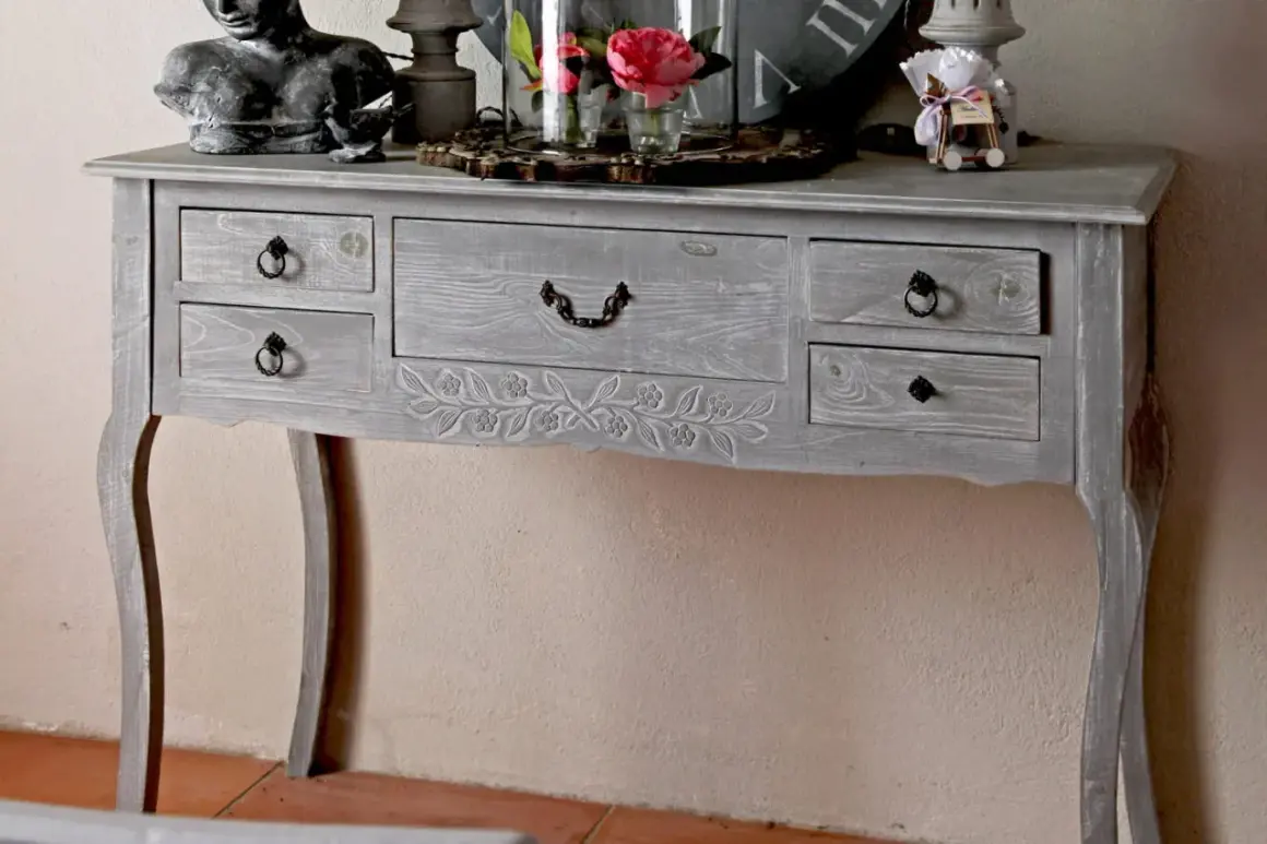Console grise patinée, ornée de motifs floraux sculptés. Ce meuble à tiroirs, idéal pour un intérieur cosy, invite à y poser des objets décoratifs.