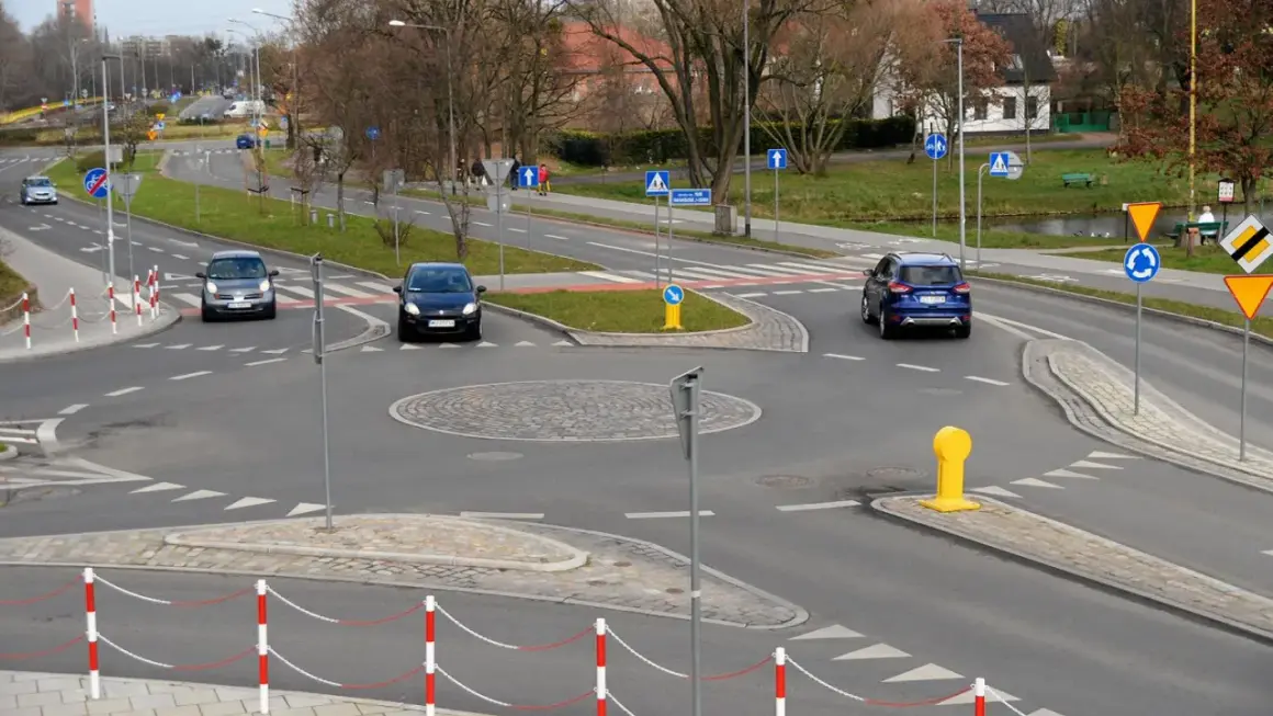 Samochody zbliżają się do ronda. Widoczny jest znak informujący o ruchu okrężnym, czyli kierunkowskaz na rondzie.