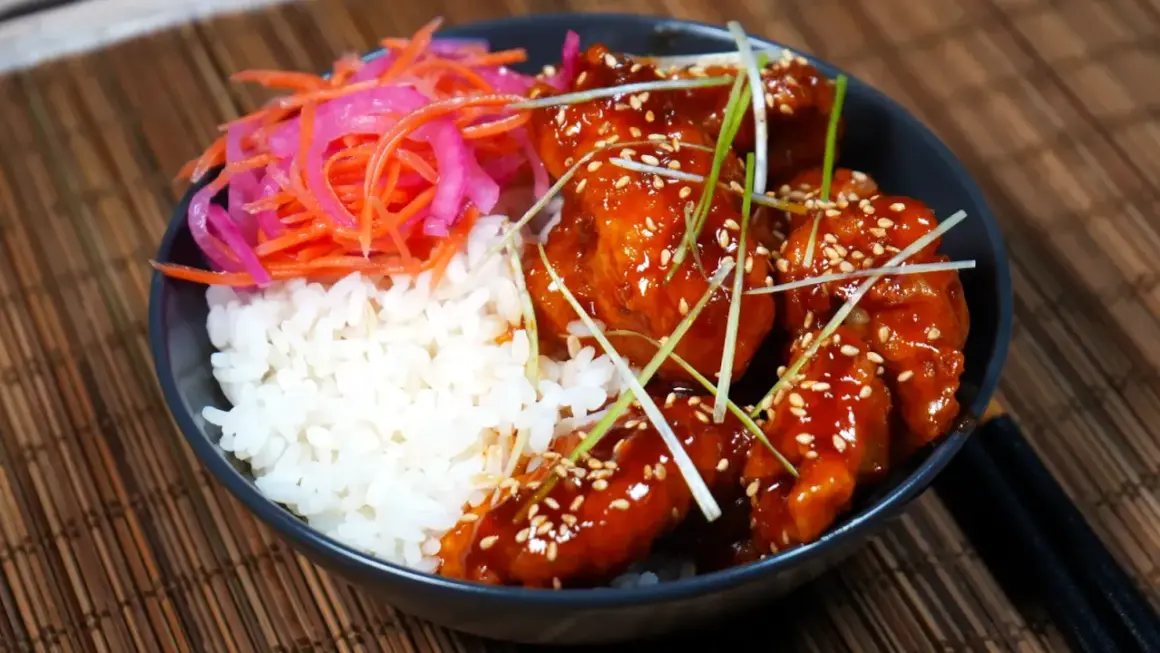 En skål med ris, krispigt koreanskt friterad kyckling i en söt och kryddig sås, toppad med sesamfrön och salladslök. Serveras med en sida av picklad morot och rödlök.