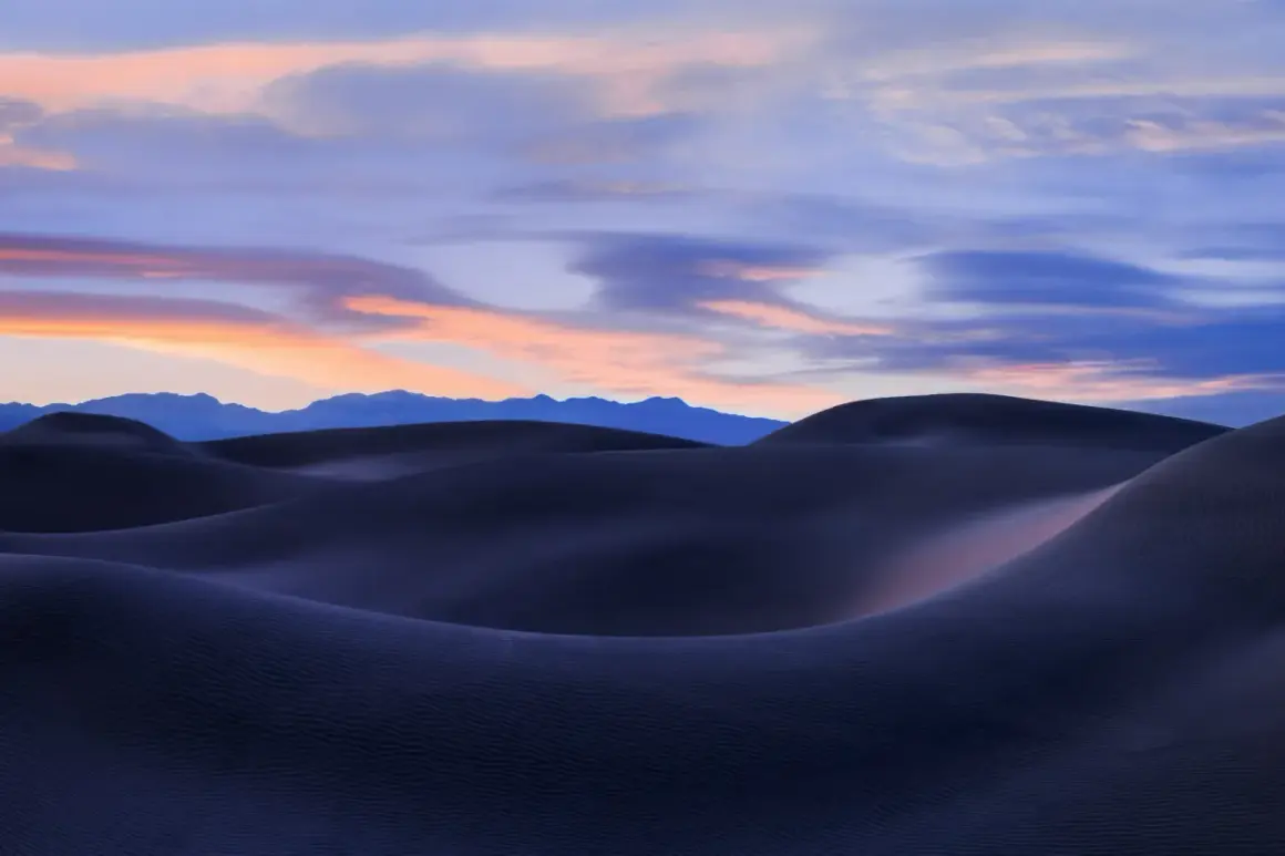 Landscape photography sunset over dark sand dunes, with soft pink and blue clouds painting the sky above distant mountains.