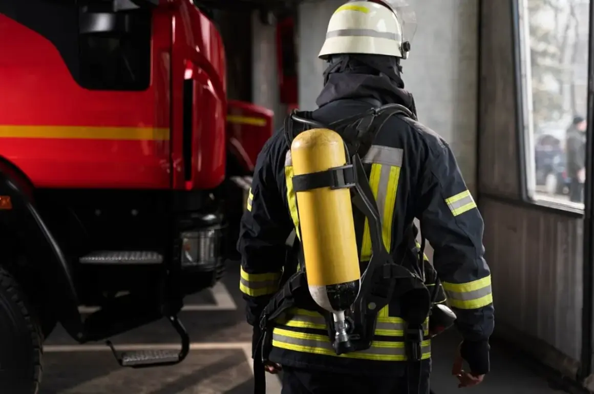 Ein Feuerwehrmann in voller Montur mit Atemschutzgerät steht bereit bei der Feuerwehr. Links im Bild ist ein Teil eines roten Feuerwehrautos zu sehen.
