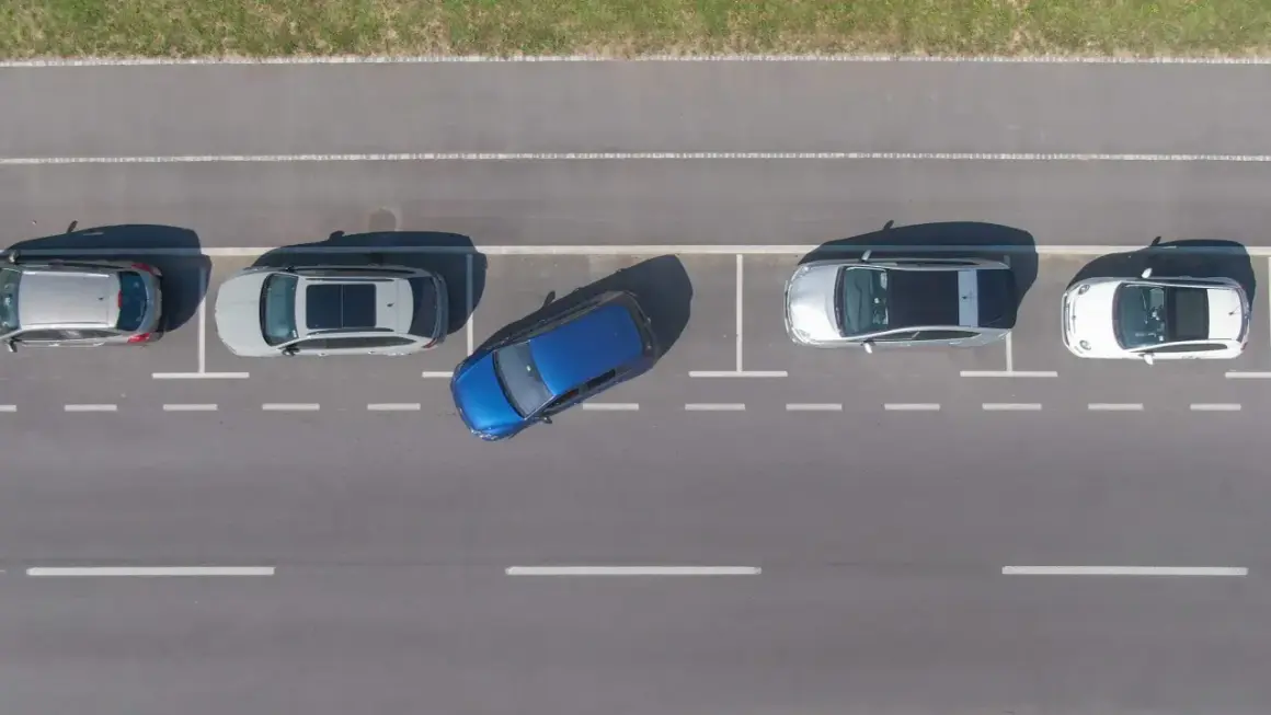 Niebieski samochód manewruje, ucząc się jak parkować równolegle między innymi autami na parkingu.
