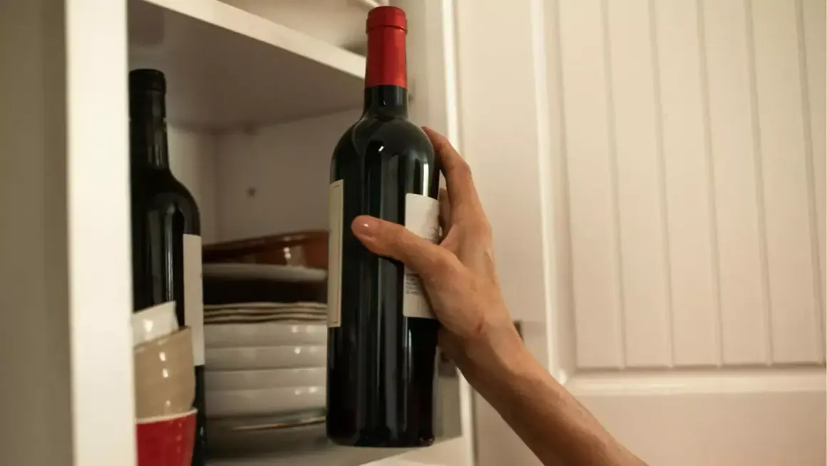 Dłoń sięga po butelkę bordeaux wina z półki, obok stosu talerzy i miseczek.