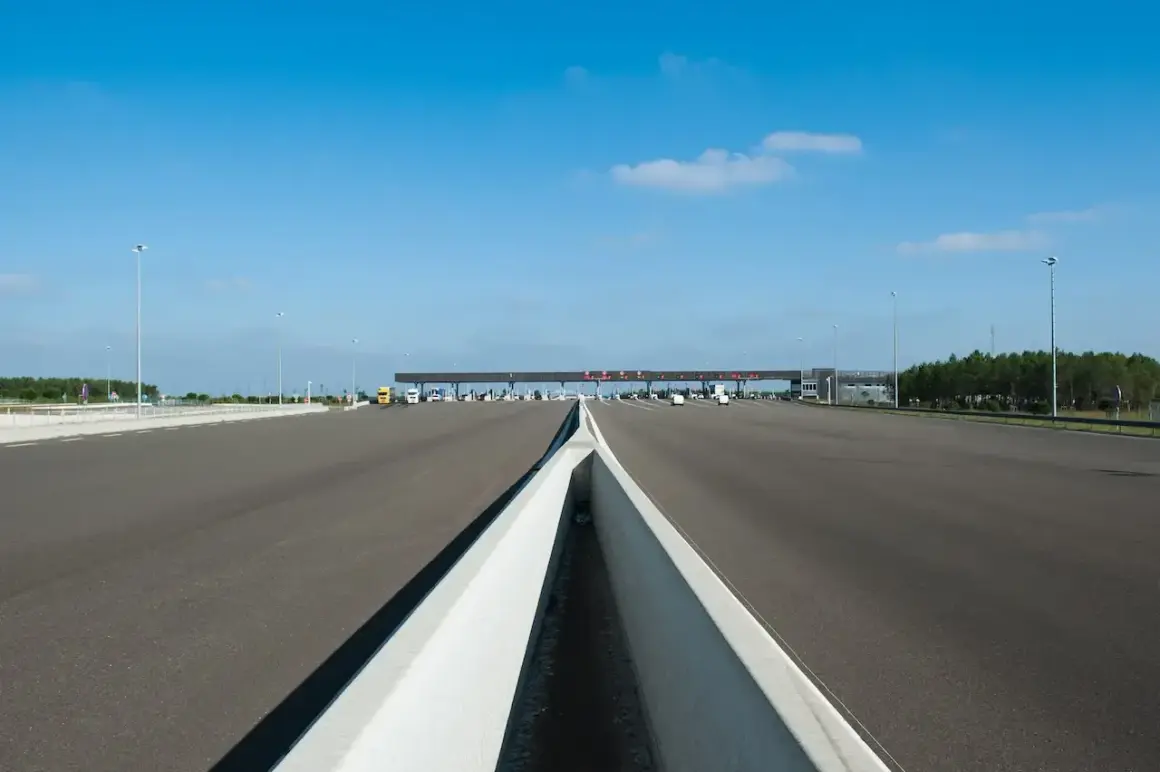 Péage autoroutier sous un ciel bleu. La route s'étend vers une barrière de péage avec plusieurs voies. Un camion jaune est visible.