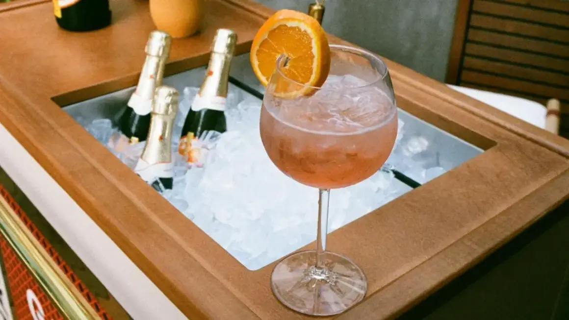 Orzeźwiający drink z prosecco i colą, ozdobiony plasterkiem pomarańczy, obok schłodzonych butelek.