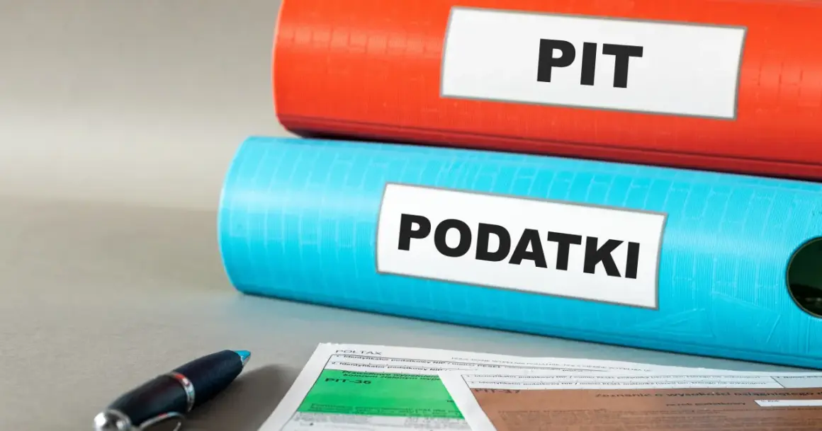 Dwa segregatory, jeden czerwony z napisem "PIT", drugi niebieski z napisem "PODATKI", leżą na biurku obok formularzy podatkowych i długopisu.
