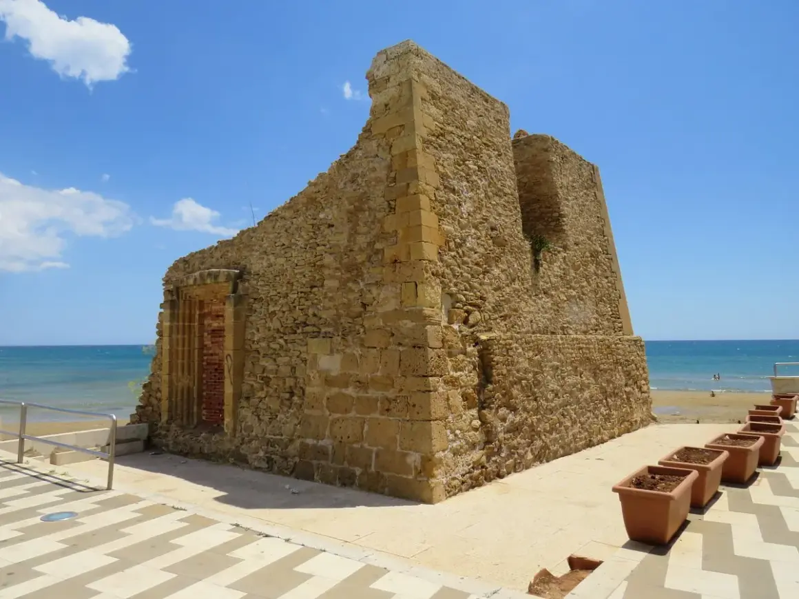 Antica torre di pietra sulla costa, con il mare azzurro sullo sfondo. Vicino, una fila di vasi di terracotta, come tre fontane in attesa.