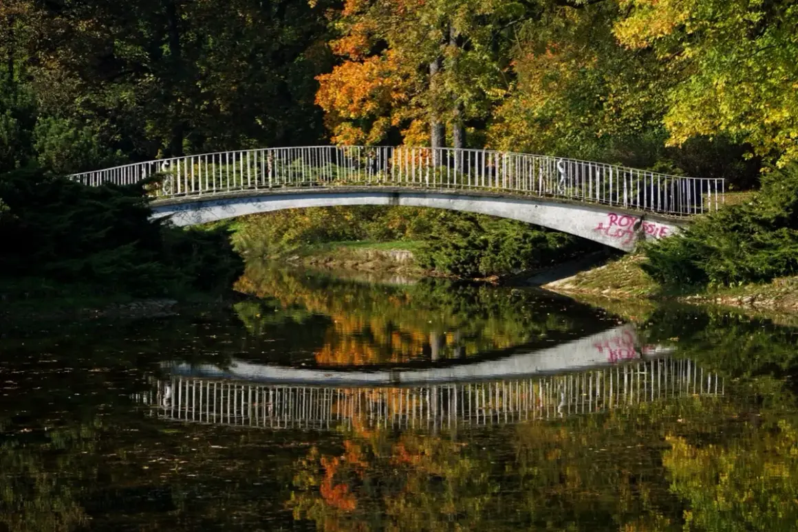 Mostek nad spokojną rzeką, odbijający jesienne barwy drzew. W oddali widać ludzi spacerujących po moście.