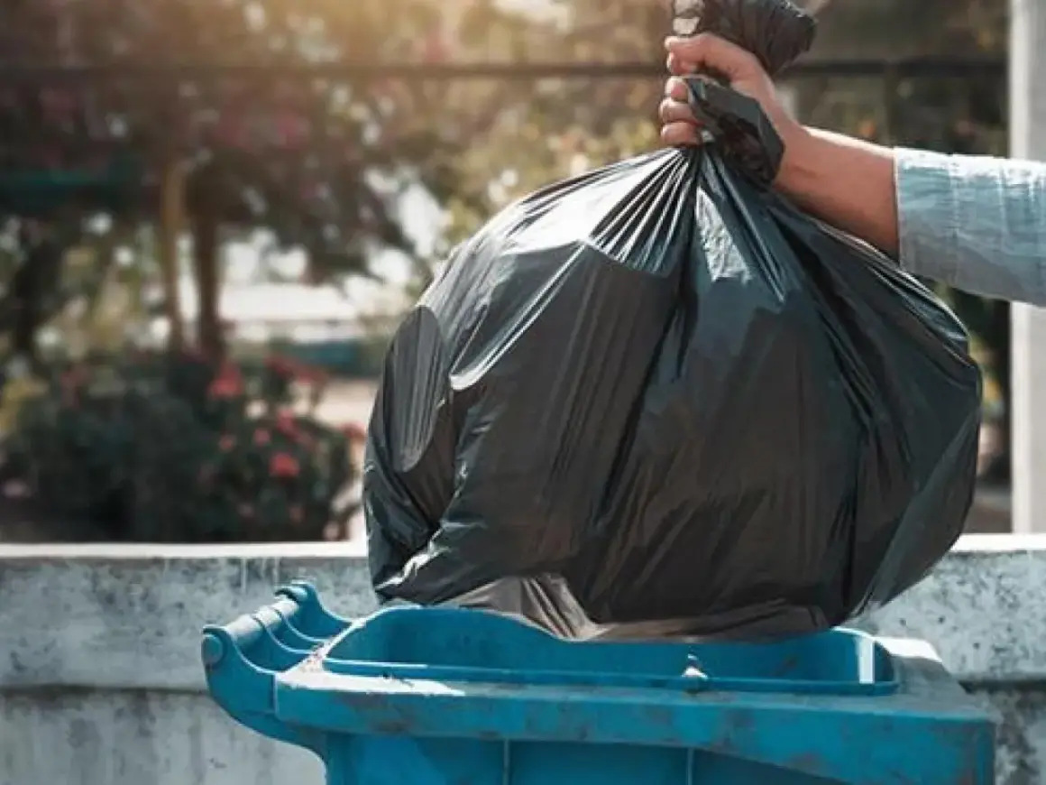 Mano depositando bolsa de basura en contenedor azul. ¿Quién paga la tasa de basura en un alquiler?