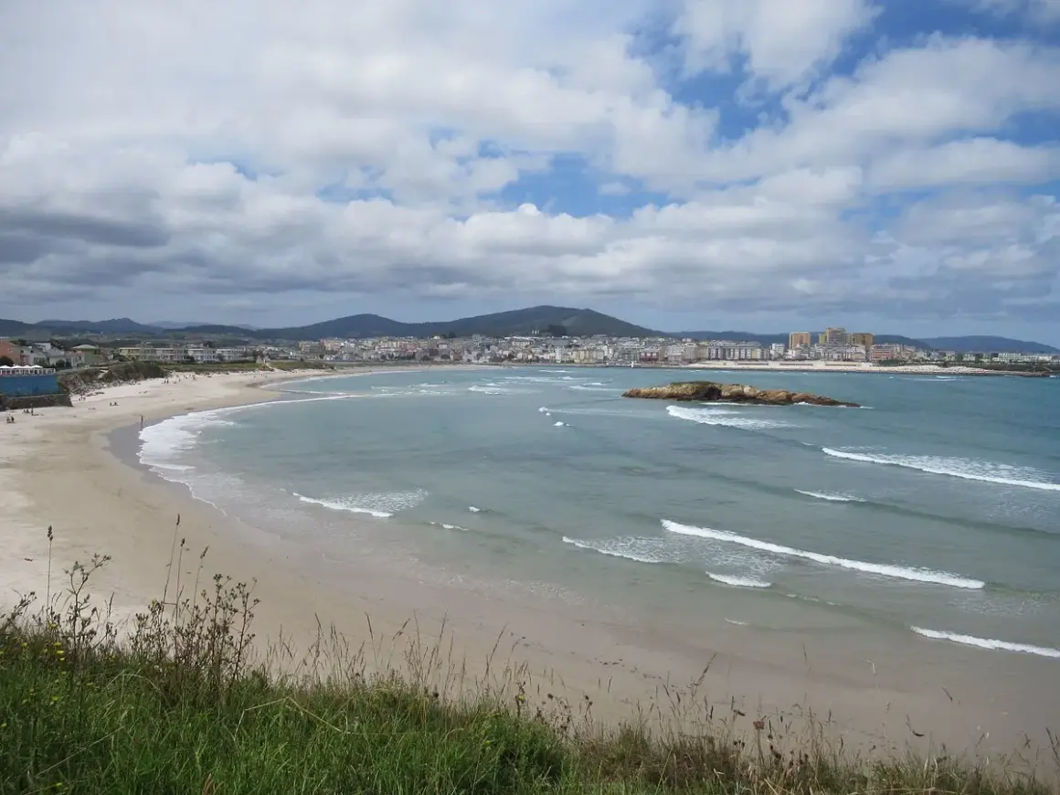 Amplia vista de la playa de Altar Barreiros con arena dorada, mar azul y una ciudad costera al fondo.