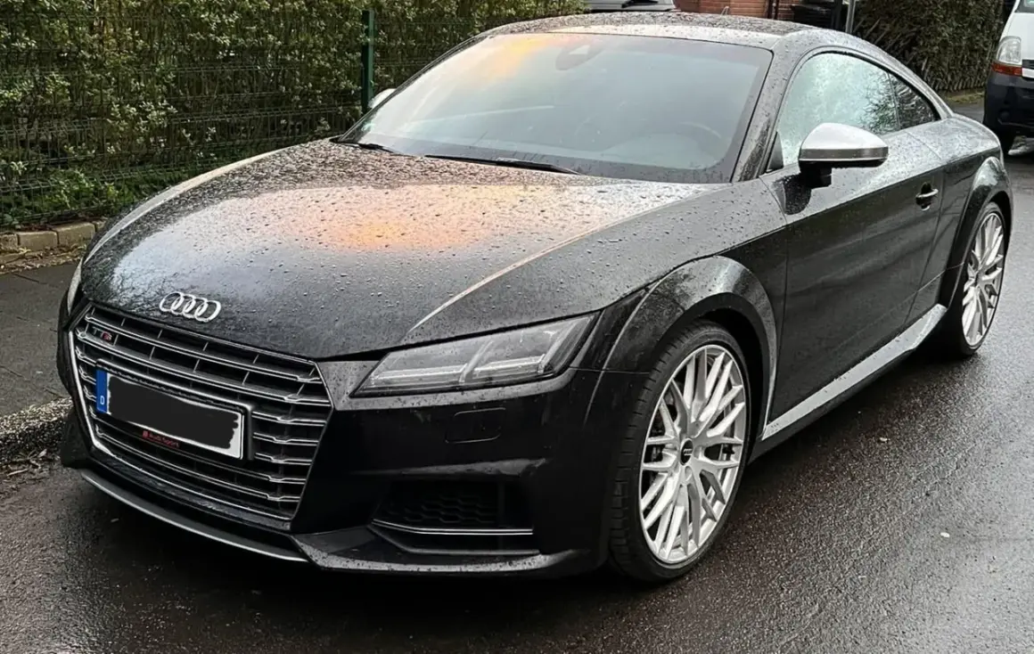 Negro Audi TT RS mojado por la lluvia, con reflejos naranjas en el capó. El coche está listo para la etapa 1 de su viaje.