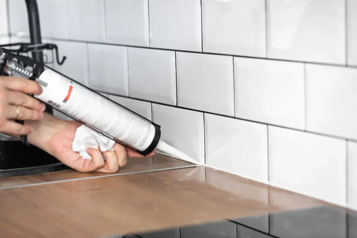 Application d'un joint d'étanchéité blanc sur un plan de travail en bois, près de carrelages muraux blancs. Le temps de séchage de la colle carrelage flex est une préoccupation pour une finition parfaite.