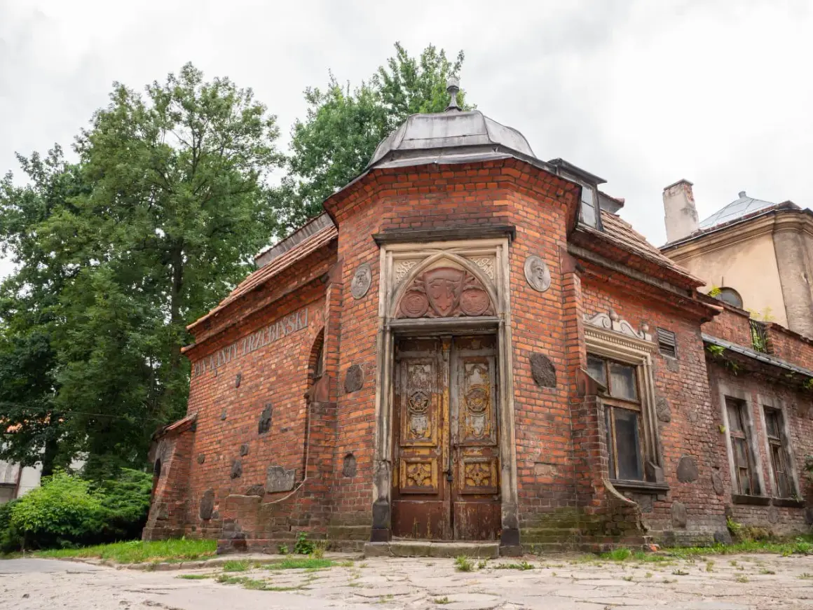 Stary ceglany budynek z ozdobnymi drzwiami i medalionami, otoczony zielenią.