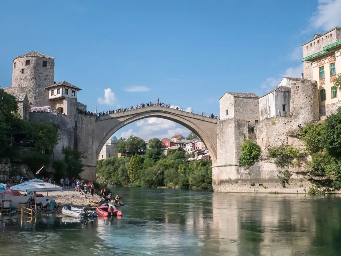 Mostar, Bośnia i Hercegowina: Słynny Stary Most z kamienia, tłumy ludzi na moście i brzegu, łodzie na rzece Neretwie.