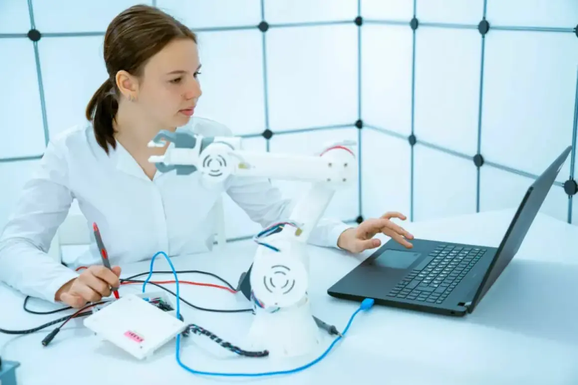 Junge Frau mit einem Roboterarm und Laptop, die an einem Projekt für ihr Robotik Studium arbeitet.