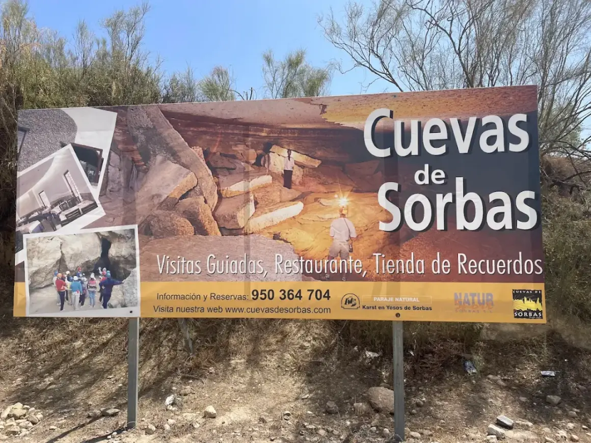 Cartel anunciando las Cuevas de Sorbas, con imágenes de exploradores y formaciones rocosas. Ofrecen visitas guiadas, restaurante y tienda.