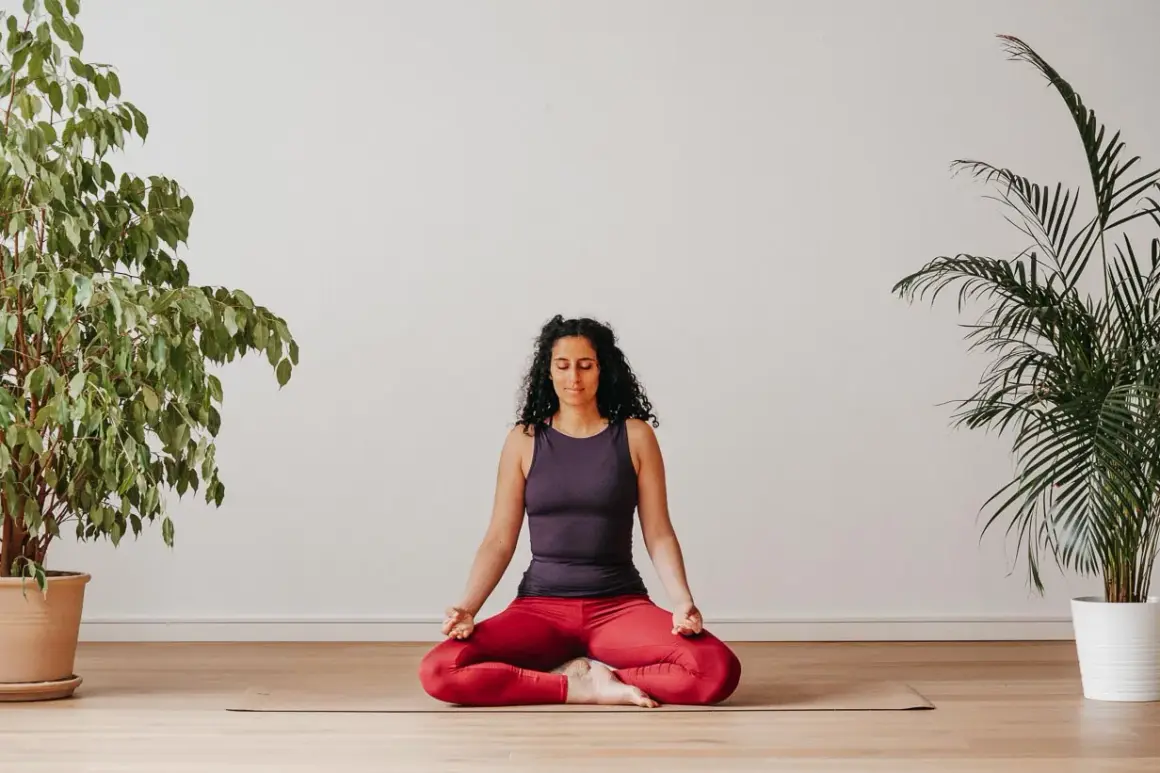 Frau praktiziert Yoga zu Hause im Lotussitz, umgeben von Pflanzen.