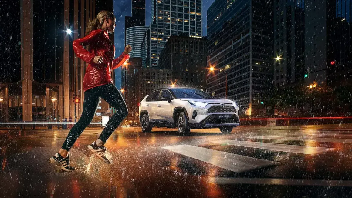 Kobieta biegnie w deszczu po mieście, obok niej jedzie biała Toyota RAV4 Hybrid. Zastanawiasz się, ile pali Toyota RAV4?