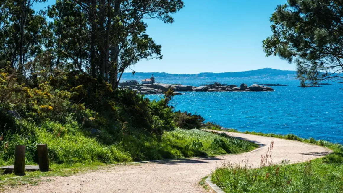 Sendero costero en la Illa de Arousa con mar azul brillante, rocas y un pequeño faro.