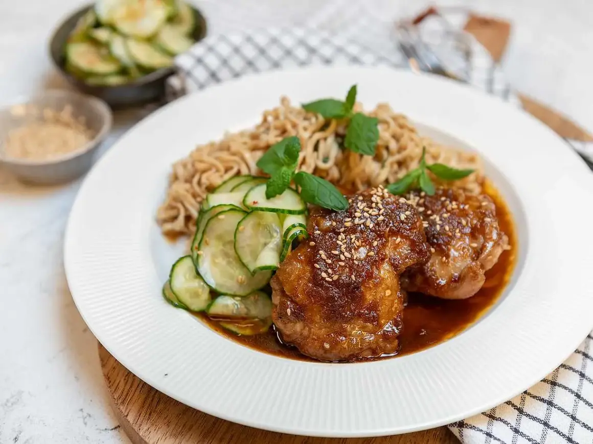Två saftiga bitar sticky chicken med sesamfrön, serverade med nudlar och gurksallad.