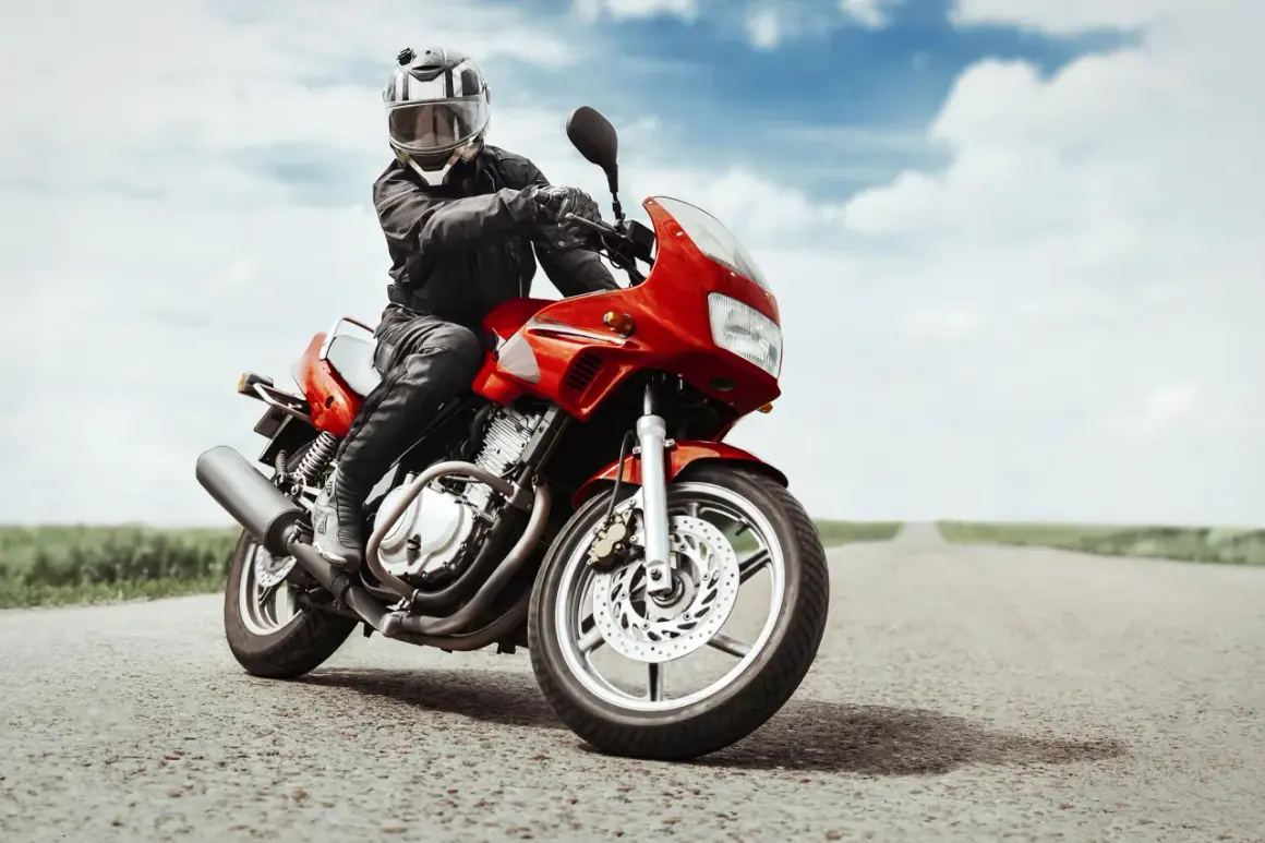 Motociclista con casco e giacca in pelle, in posizione di guida sportiva su una moto rossa su una strada vuota.