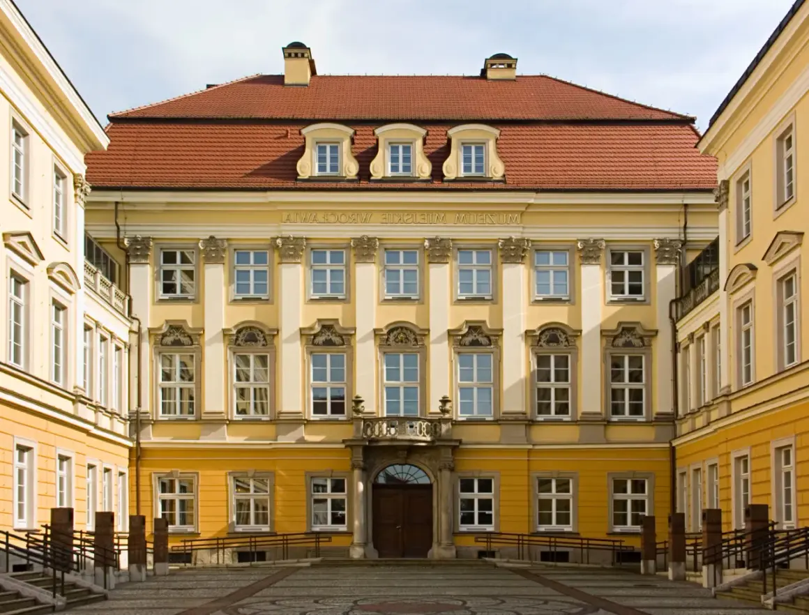 Elegancki budynek z żółtą fasadą i czerwonym dachem, ozdobiony licznymi oknami. To muzeum sztuki medalierskiej we Wrocławiu.