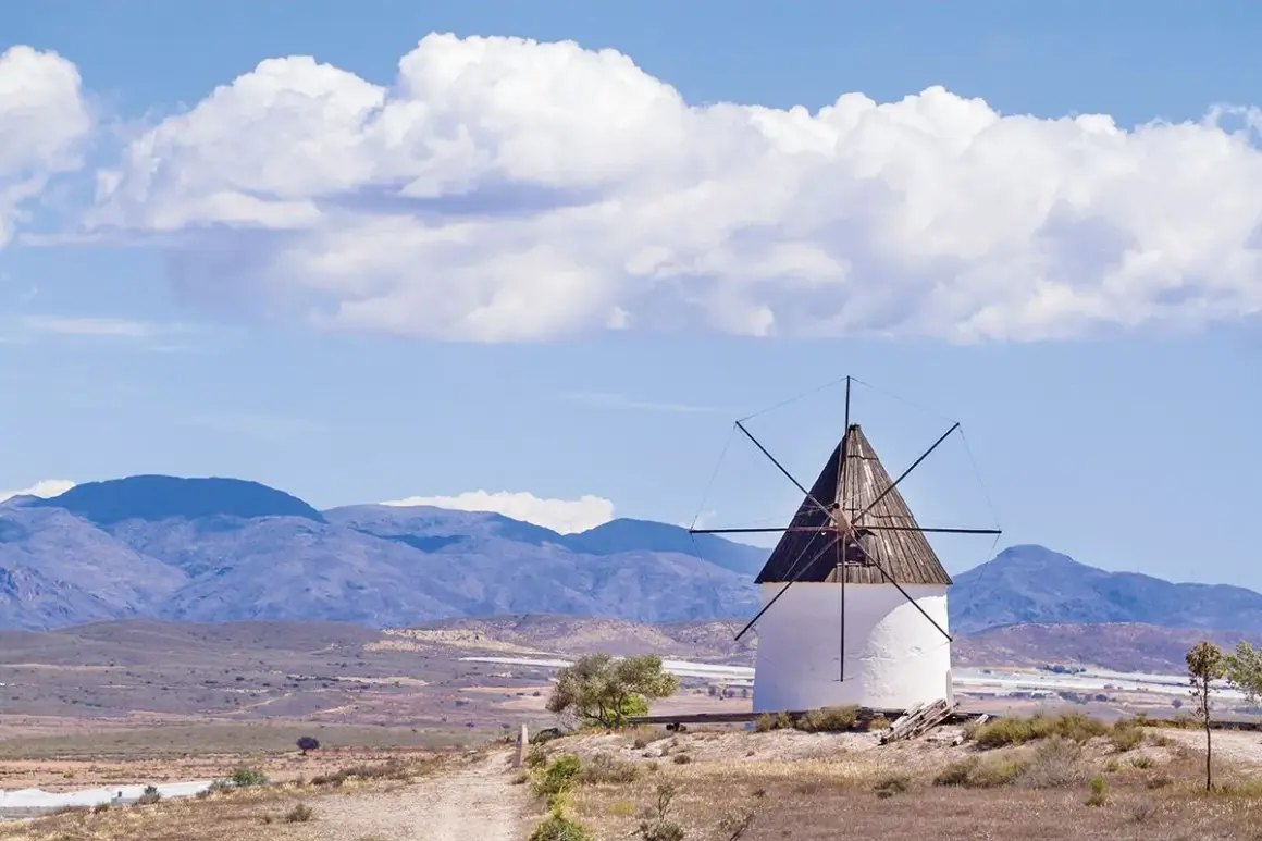 Molino blanco con aspas de madera bajo un cielo azul con nubes. Paisaje árido con montañas al fondo, como si Fernan Perez hubiera pintado este cuadro.