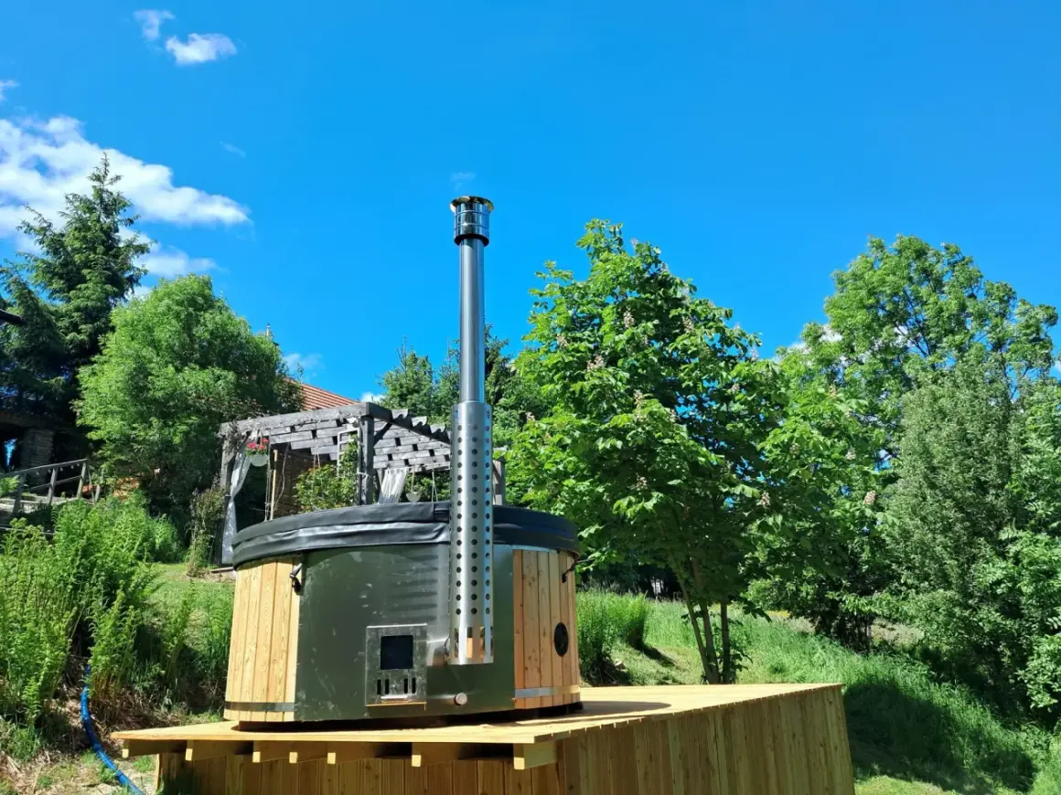 Drewniane jacuzzi w ziemi z metalowym kominem, otoczone zielenią i błękitnym niebem. Idealne miejsce na relaks.