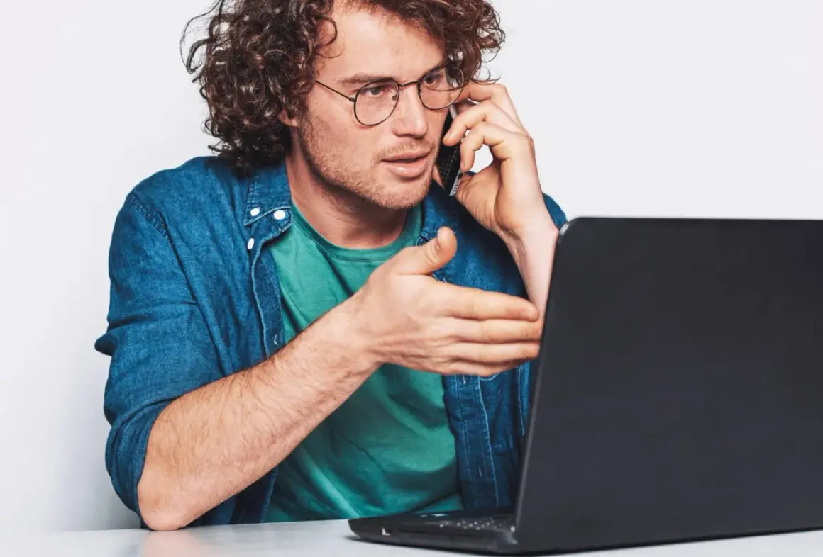 Mężczyzna w okularach rozmawia przez telefon, gestykulując w stronę laptopa. Szuka uczciwych firm oddłużeniowych, by rozwiązać swoje problemy finansowe.