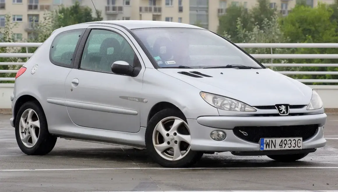 Srebrny Peugeot 206 na mokrym parkingu, z widocznym numerem rejestracyjnym WN 4933C.