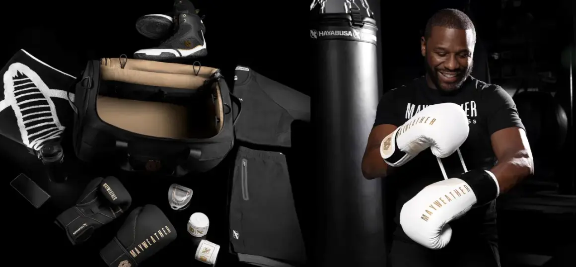 Floyd Mayweather Jr. smiles, holding a pair of white and gold Mayweather boxing gloves. The image also displays his boxing gear, including black gloves, shoes, and apparel.