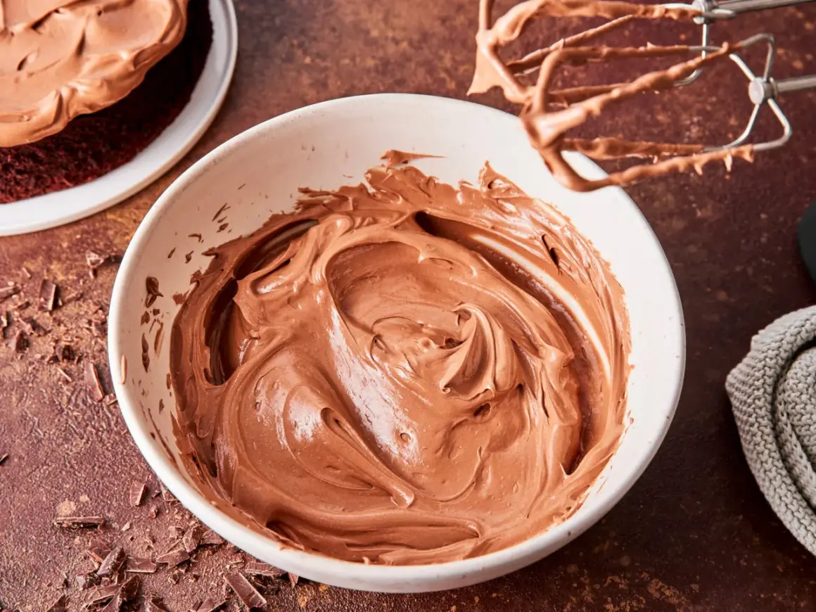 Schoko-Buttercreme in einer Schüssel, bereit zum Verzieren eines Kuchens. Ein Handmixer mit Schoko-Buttercreme hängt darüber.