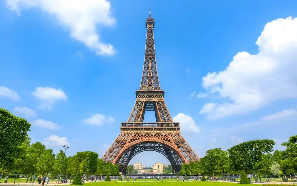 The Eiffel Tower stands tall against a bright blue sky with fluffy white clouds, surrounded by lush green trees and a manicured lawn.