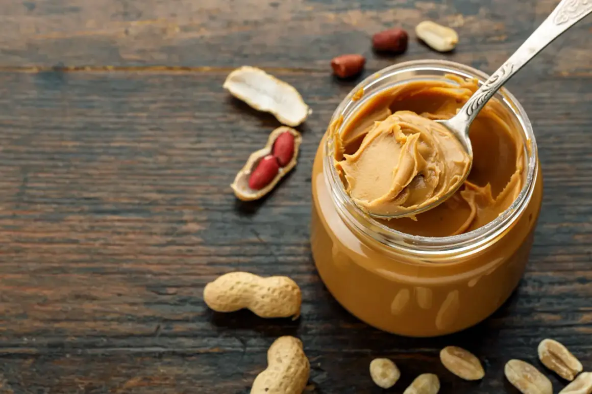 Ein Löffel cremiger Erdnussbutter aus einem Glas, umgeben von Erdnüssen. Erdnussbutter gesund und lecker!