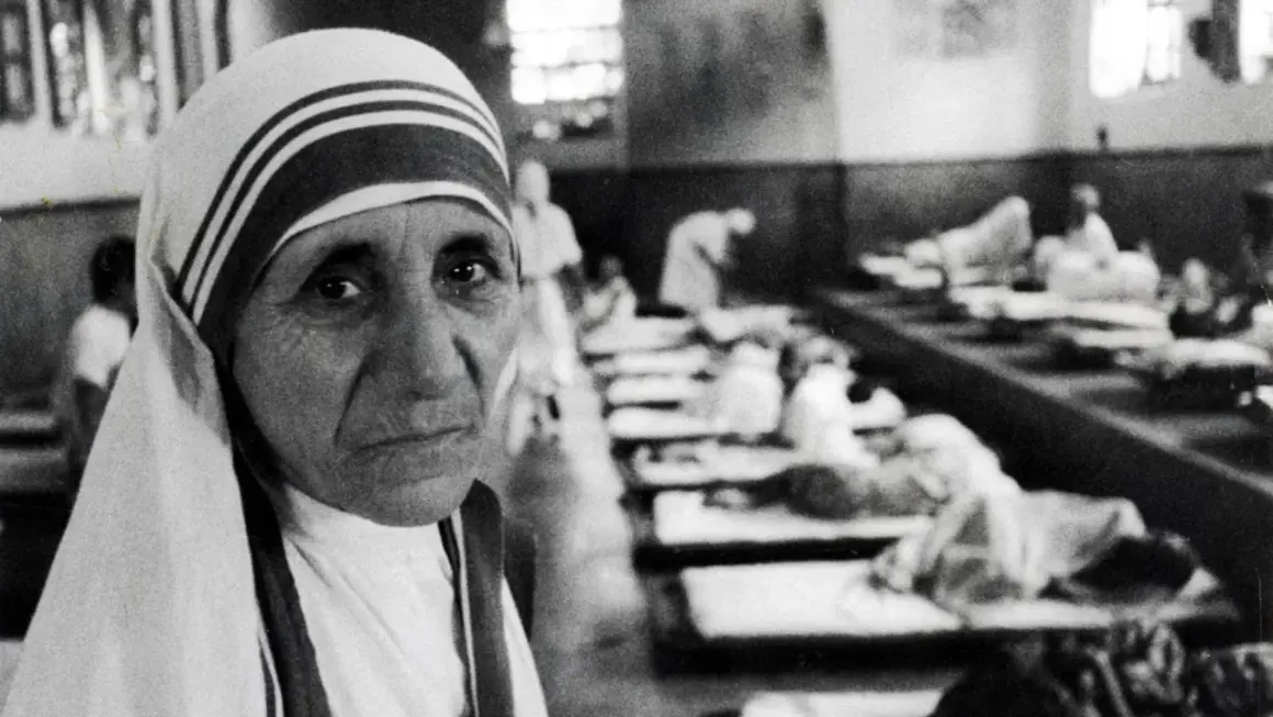 Mutter Teresa, deren Herkunft in Skopje liegt, blickt nachdenklich in die Kamera. Im Hintergrund liegen Kranke in Betten.