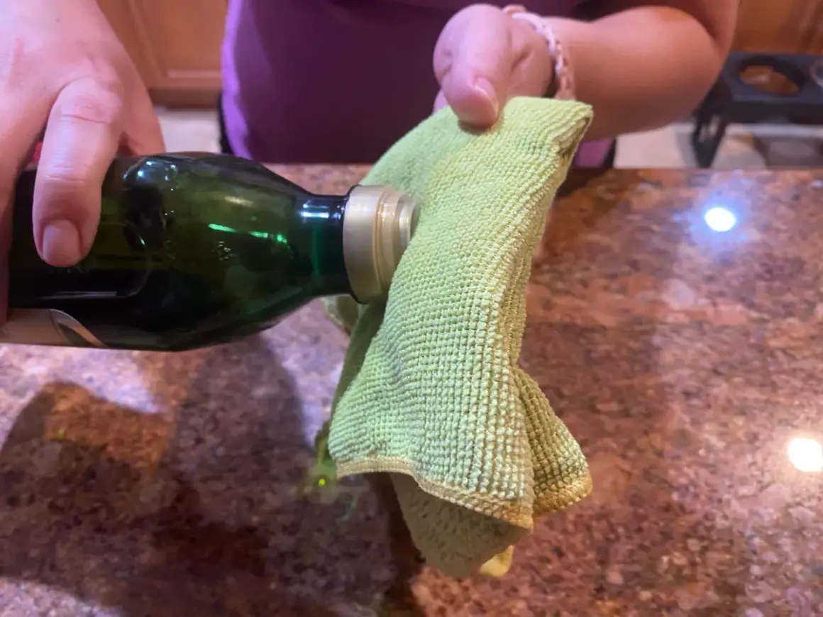 A hand pours olive oil from a green bottle onto a green microfiber cloth, suggesting it's the best home remedy to clean stainless steel.