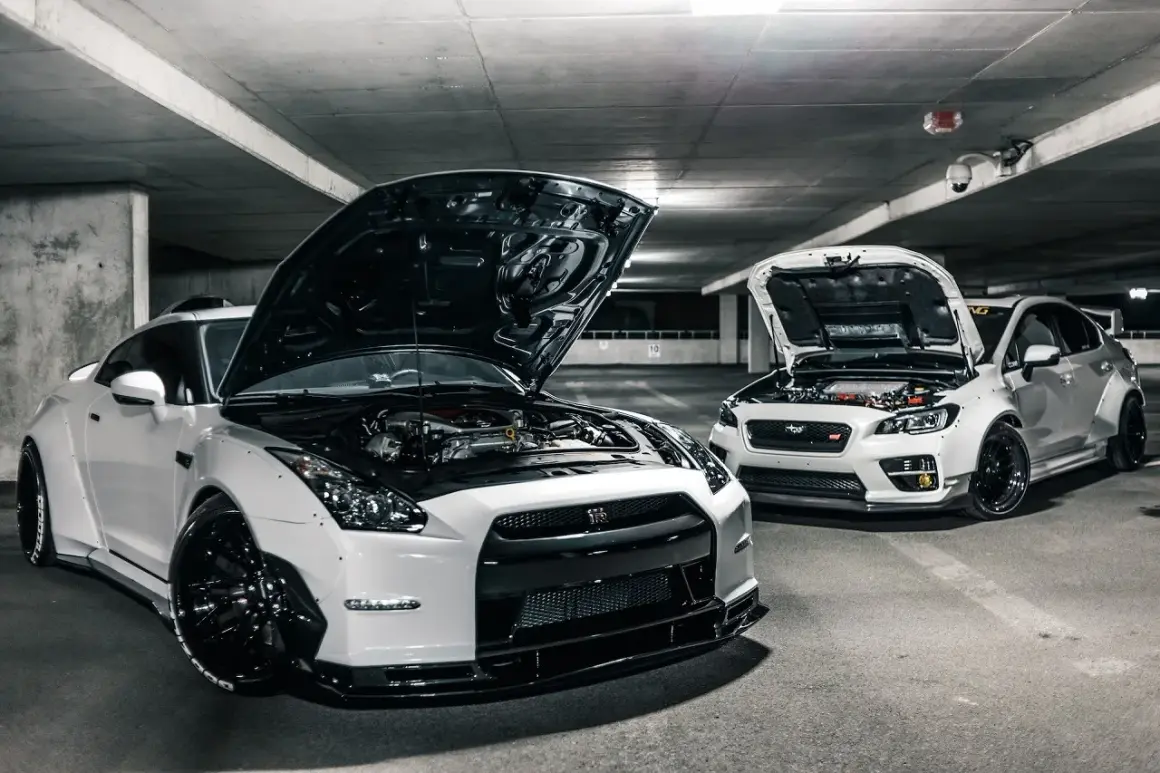 Deux voitures de sport blanches, capots ouverts, révélant leurs moteurs. Ces chevaux fiscaux, prêts à rugir, sont garés dans un parking sombre.