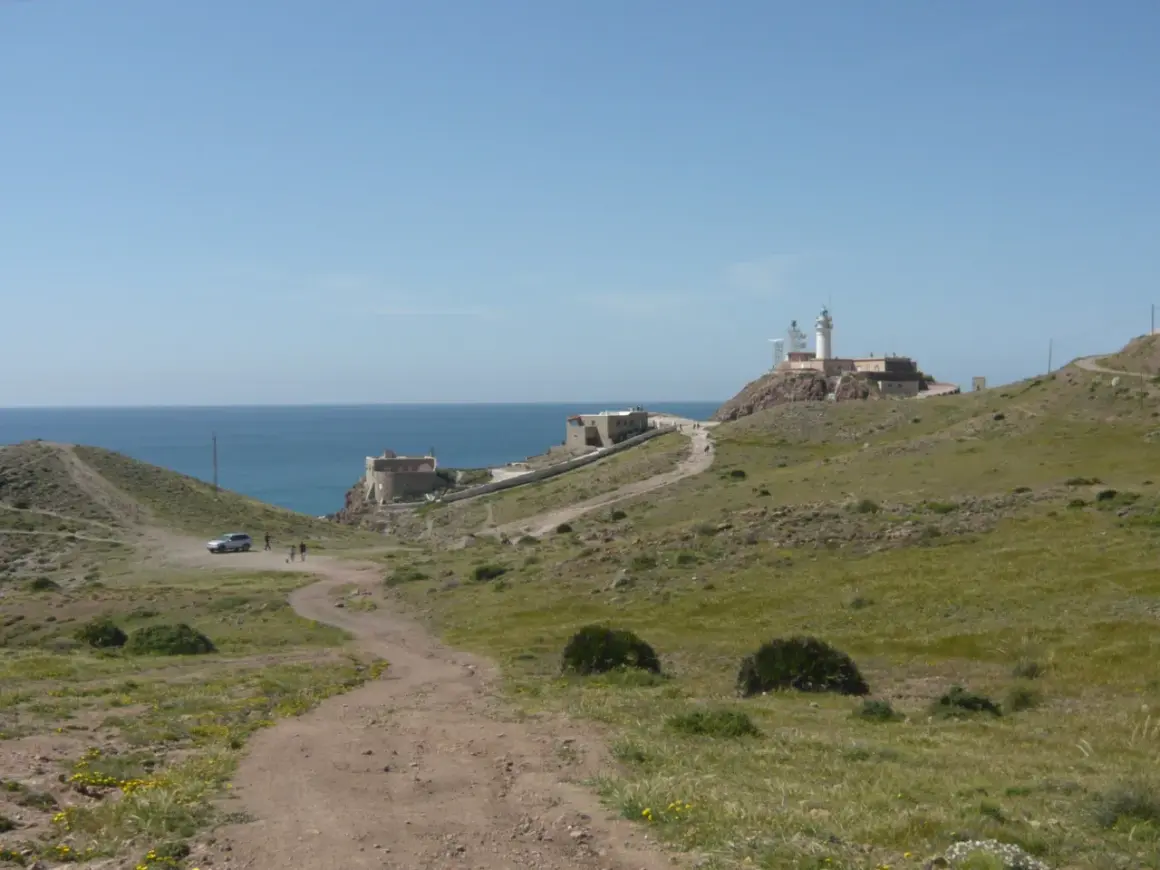 Un camino de tierra lleva al restaurante El Faro Cabo de Gata, con el mar azul y un faro en la colina.