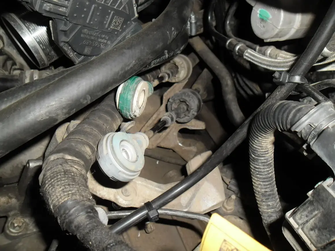 Widok na mechanizm skrzyni biegów z widocznymi linkami zmiany biegów, przewodami i elementami silnika.