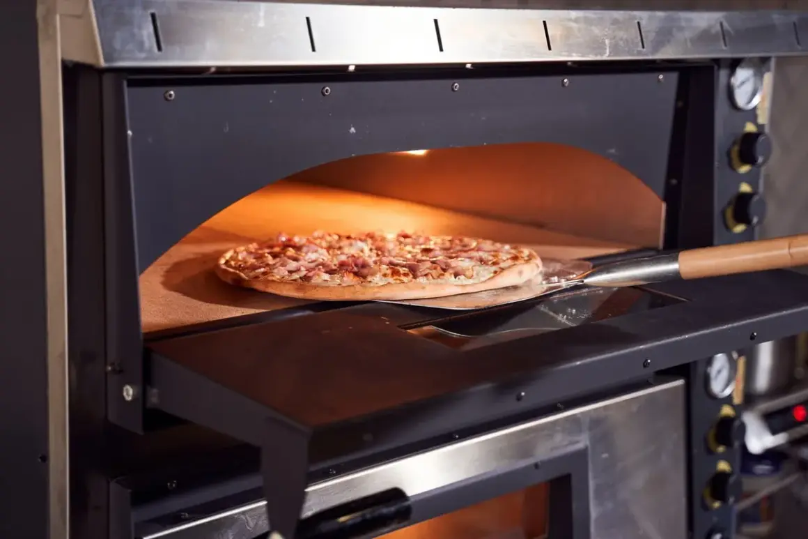 Une pizza garnie est en cours de cuisson dans un four professionnel, sur une pelle à pizza. La chaleur intense du four promet une cuisson parfaite.