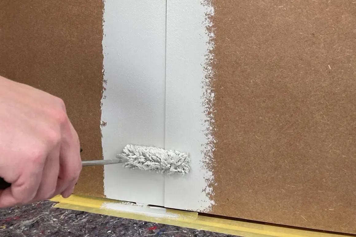 Hand streicht mit einer Farbrolle weiße Farbe auf folierte Paneele. Der Boden ist mit Malervlies und Klebeband geschützt.