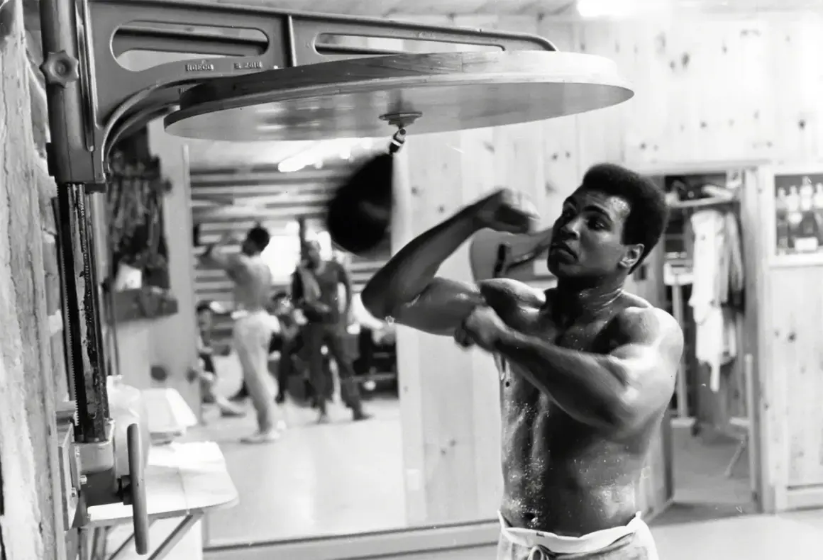 Muhammad Ali trains with a speed bag, sweat glistening on his skin, a testament to the dedication behind his many achievements.