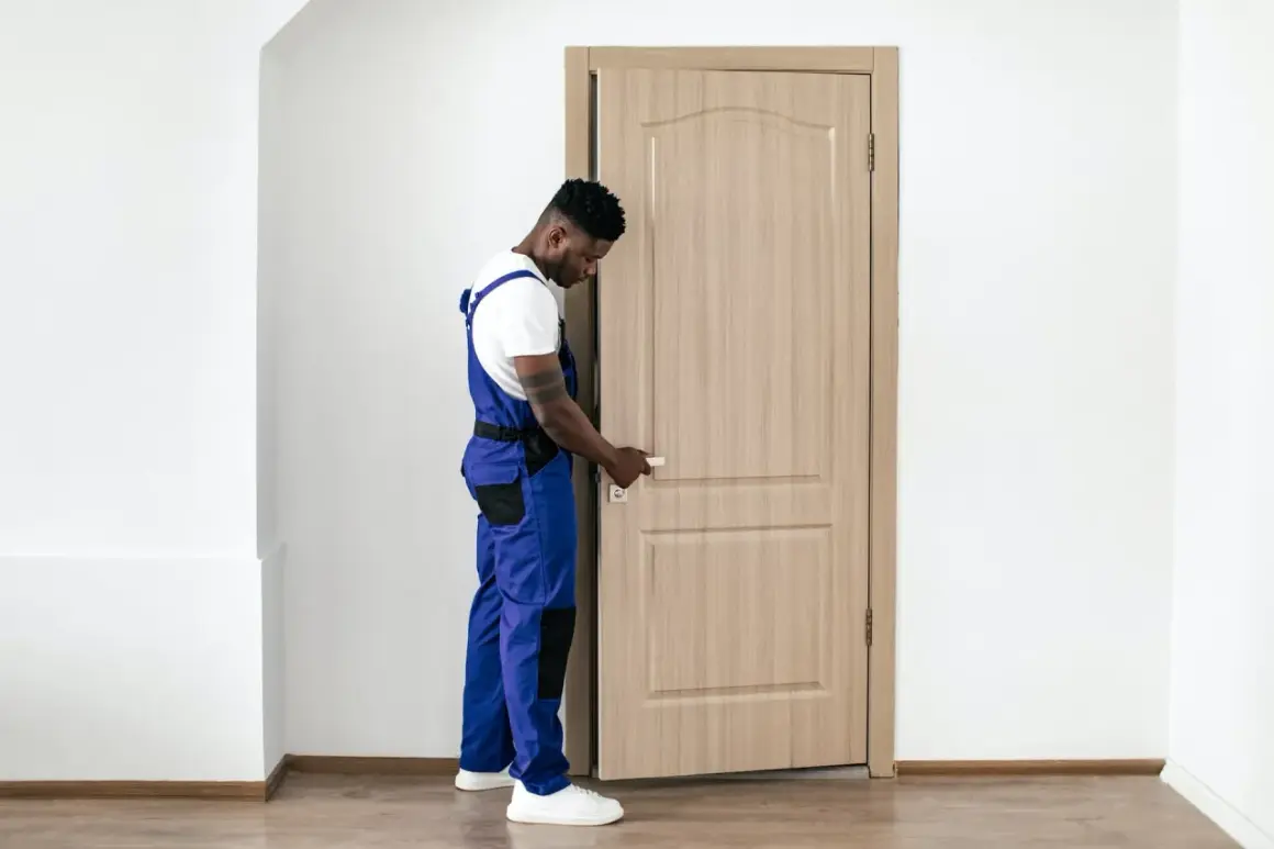 Un ouvrier en salopette bleue manipule la poignée d'une porte intérieure en bois clair, prêt à la changer.