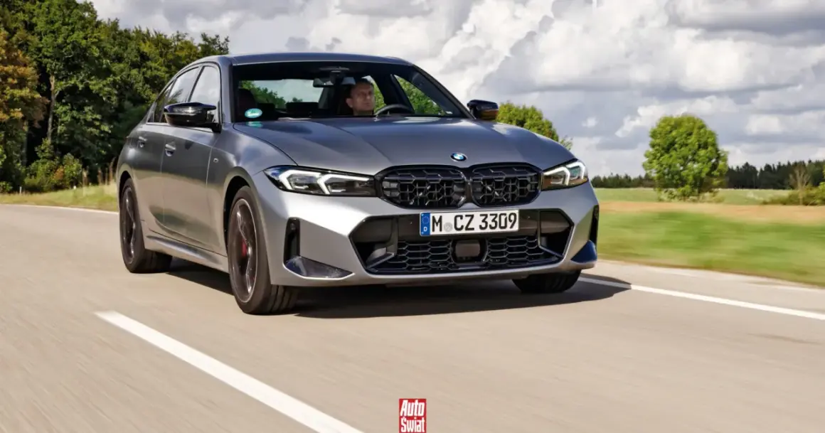 Szare BMW 320d jedzie po drodze, za kierownicą siedzi mężczyzna. W tle drzewa i chmury.